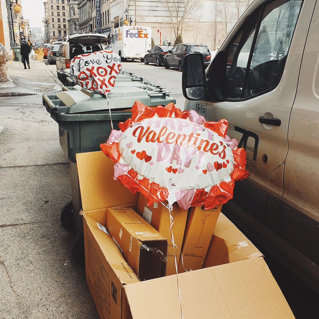 Valentine's Day balloons floating out of an open cardboard box on a New York City sidewalk beside a delivery van