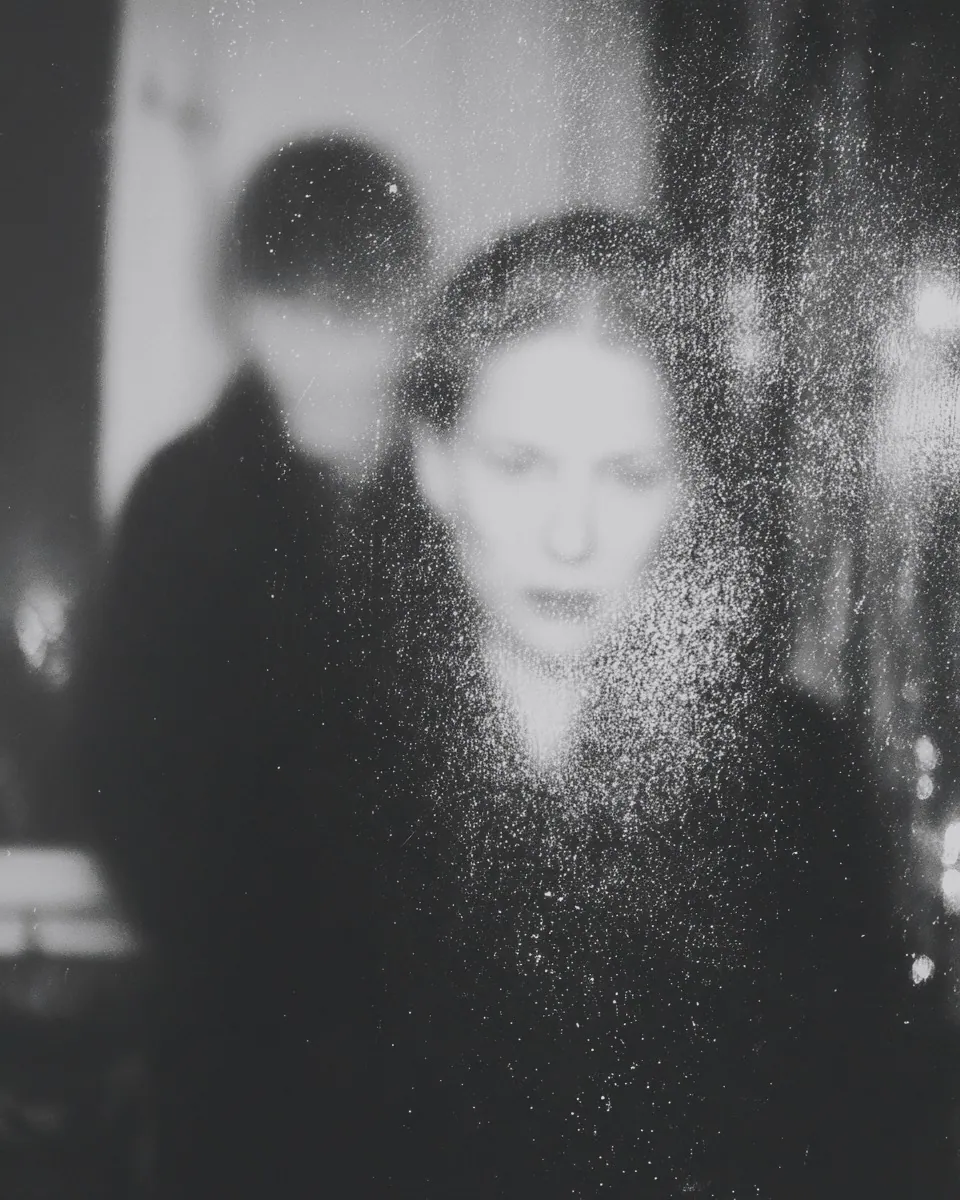 A woman with a speckled, dusty face stands in front while a darker blurred figure looms behind her.