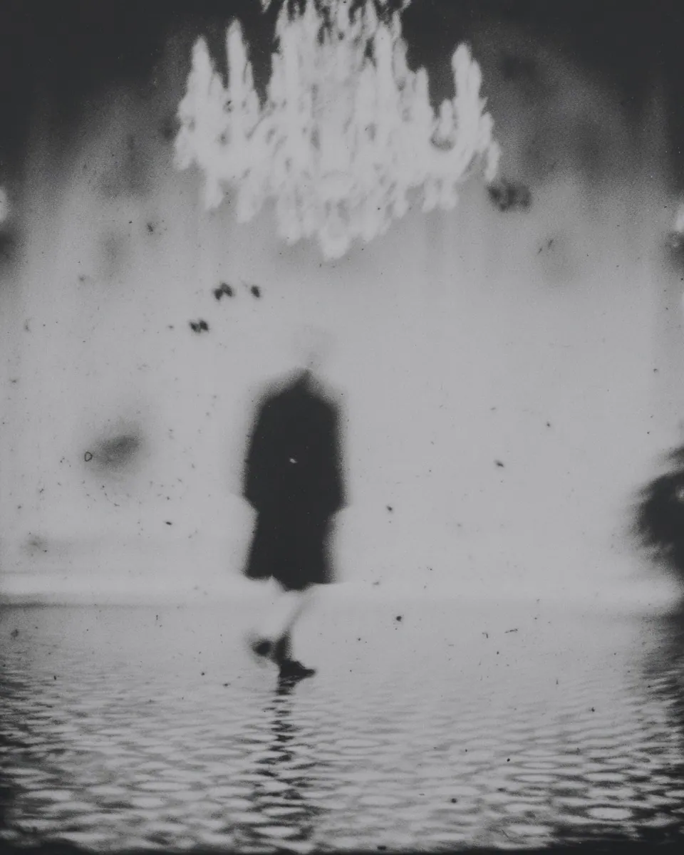 A dark figure walks across a flooded floor beneath a glowing chandelier.