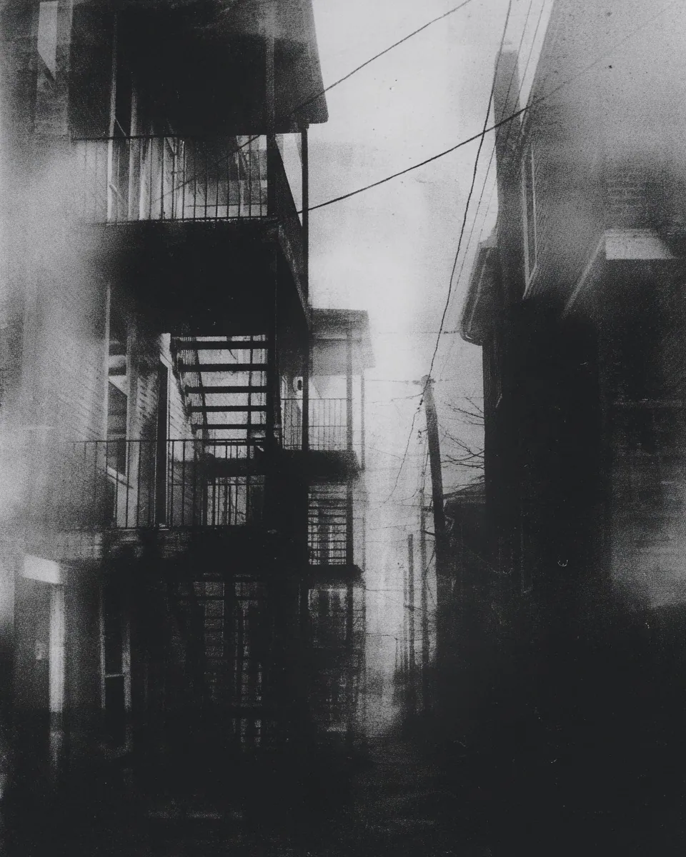 A narrow back alley of brick apartments with metal fire escapes and tangled overhead wires, washed in haze.