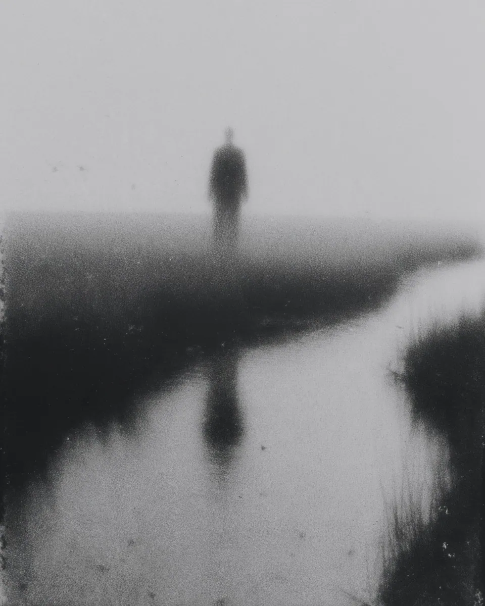 A dark figure stands at the end of a winding creek in the fog, mirrored in the water.