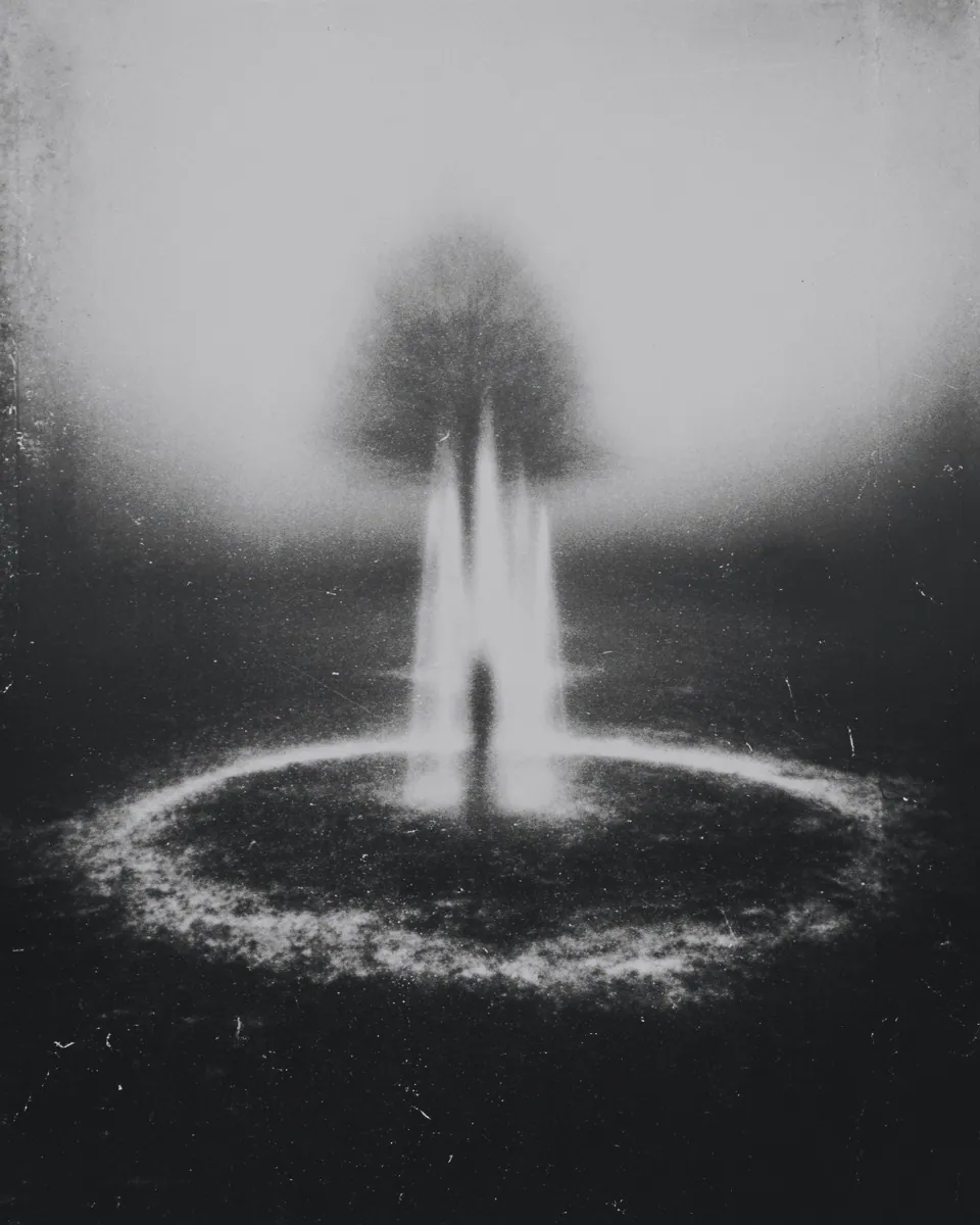 A lone figure stands inside a fountain at the centre of a pale circle, a tree blurred in the fog behind.