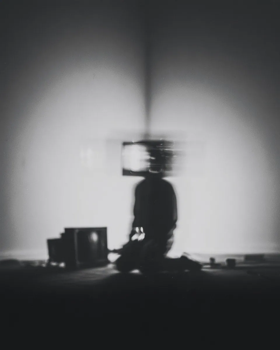 A blurred figure kneels beside old televisions in an empty room, the head replaced by a glowing screen.