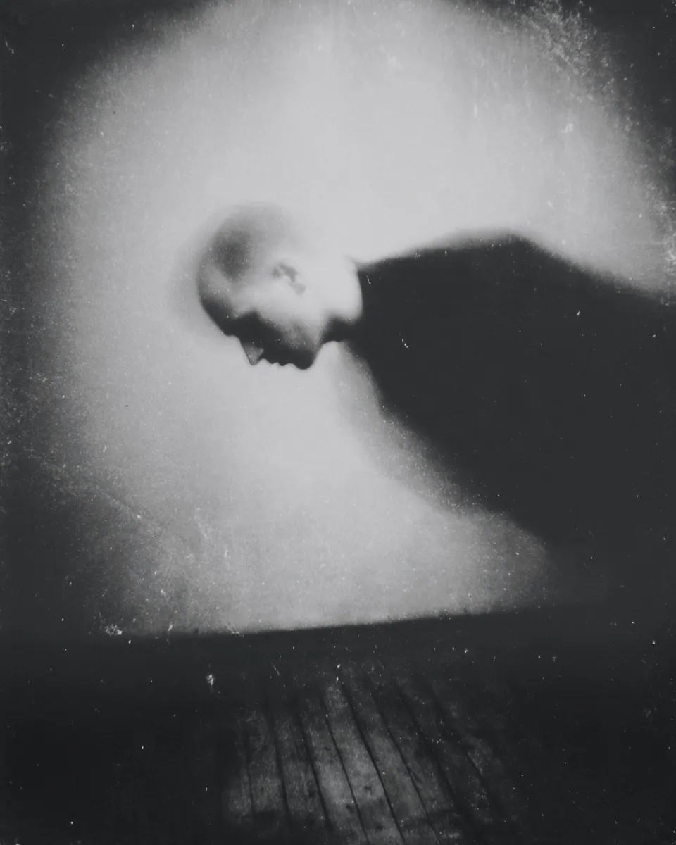 A bald man bends forward, his body dissolving into a dark blur against a pale wall.