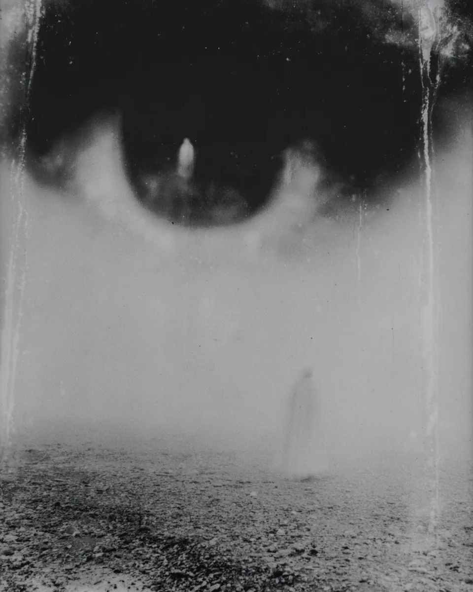 A huge dark eye looks down from the sky over a faint pale figure on a gravel plain.