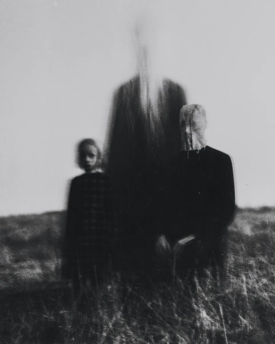A child and a masked adult sit in tall grass, a tall dark shape looming between them.