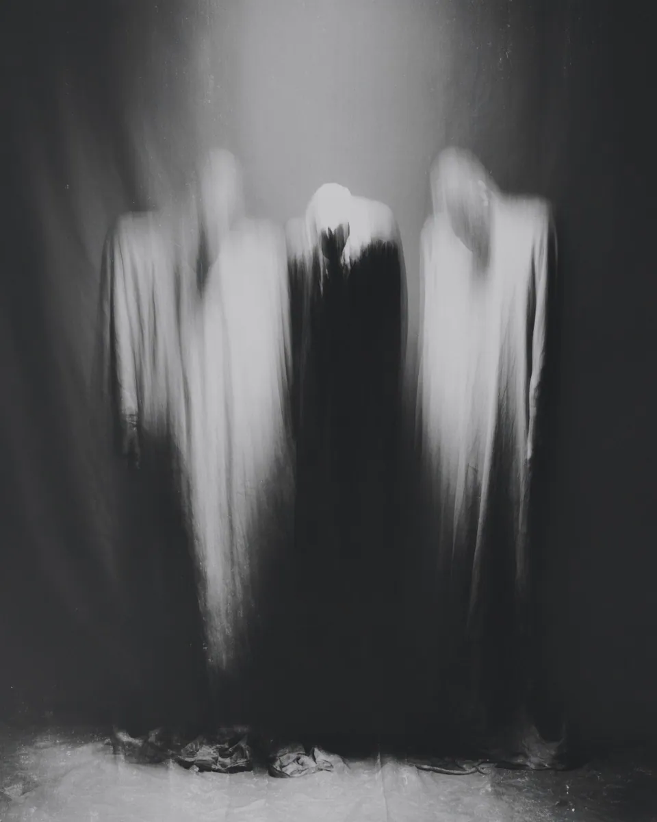 Three draped, ghostly figures in white and black stand together against a dark curtain.