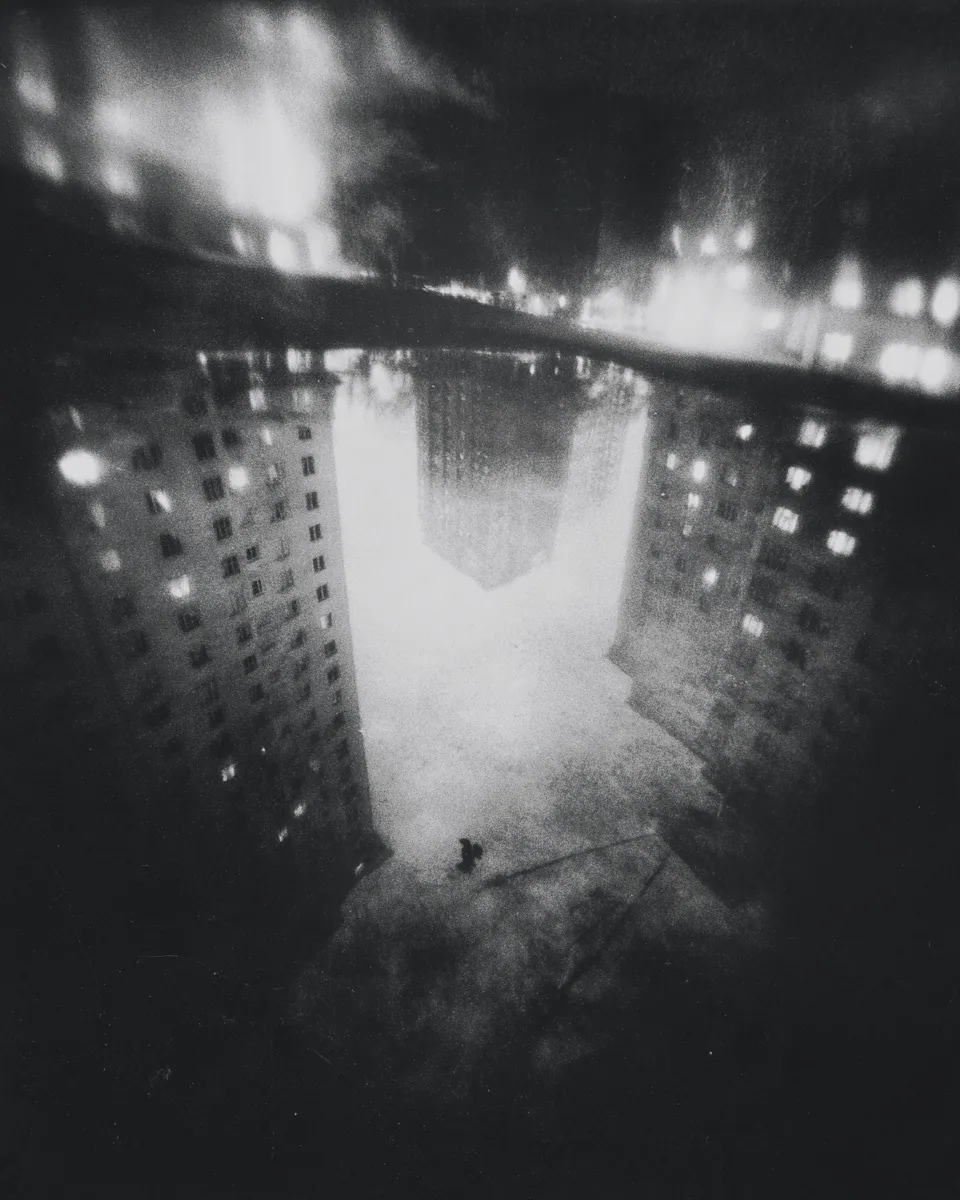 Towers with lit windows reflect in a puddle, turning the street into an inverted city at night.