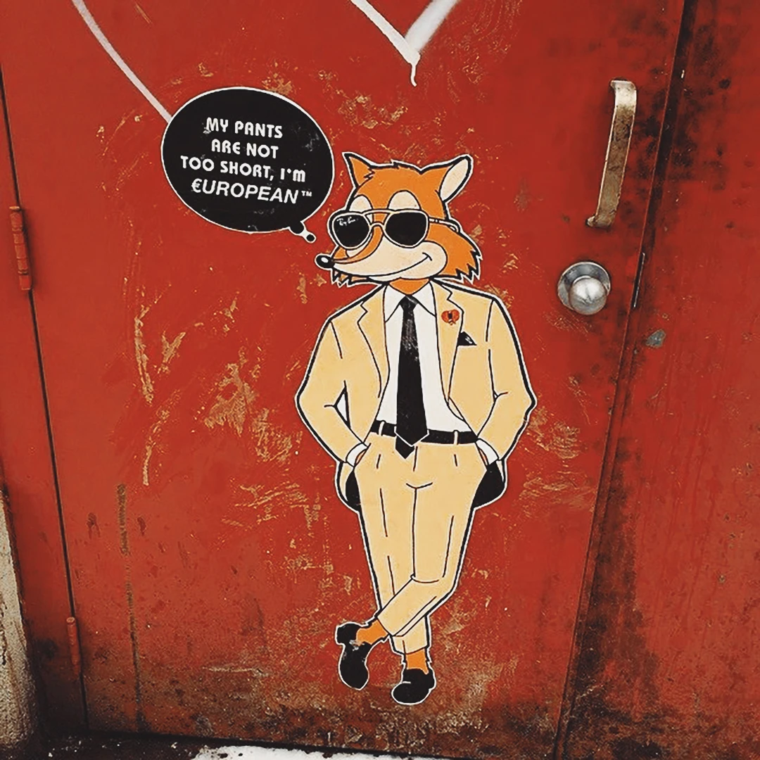Street art of a dapper fox in a beige suit on a red door, speech bubble reading 'My pants are not too short, I'm European'