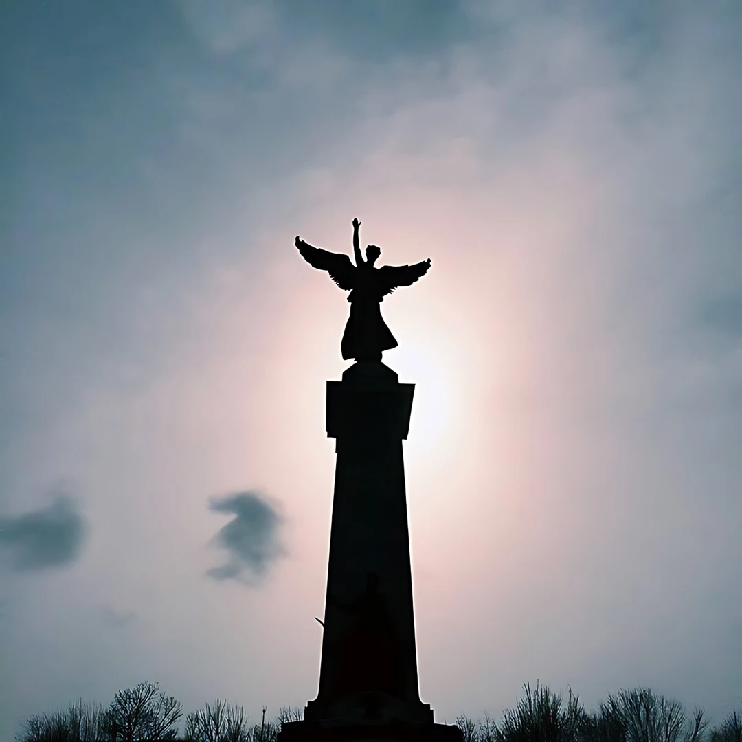 Silhouette of a winged angel statue atop a tall column, backlit by hazy sunlight breaking through clouds
