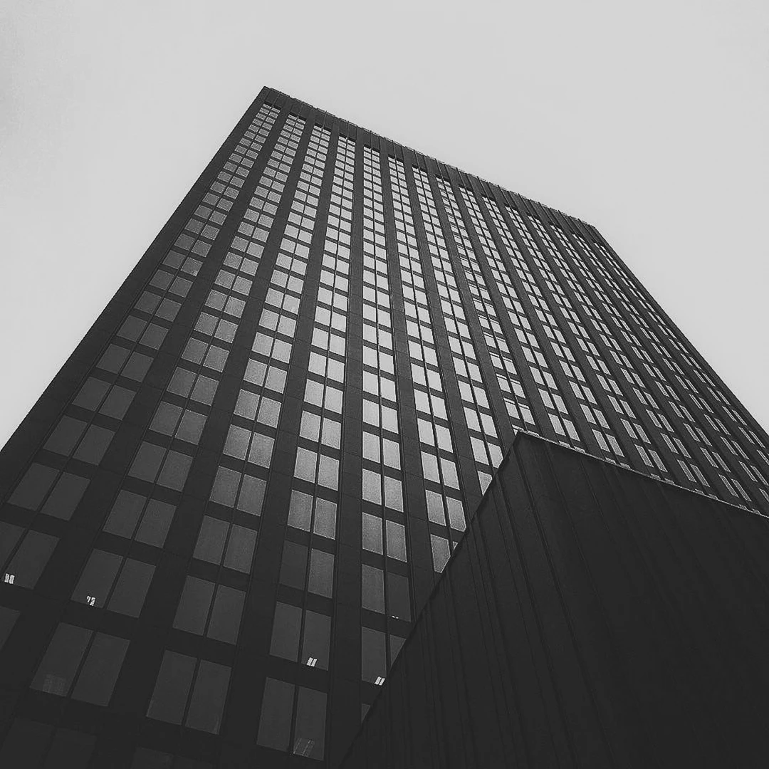 Black and white low-angle shot of a glass skyscraper's repeating window grid dissolving into a grey overcast sky