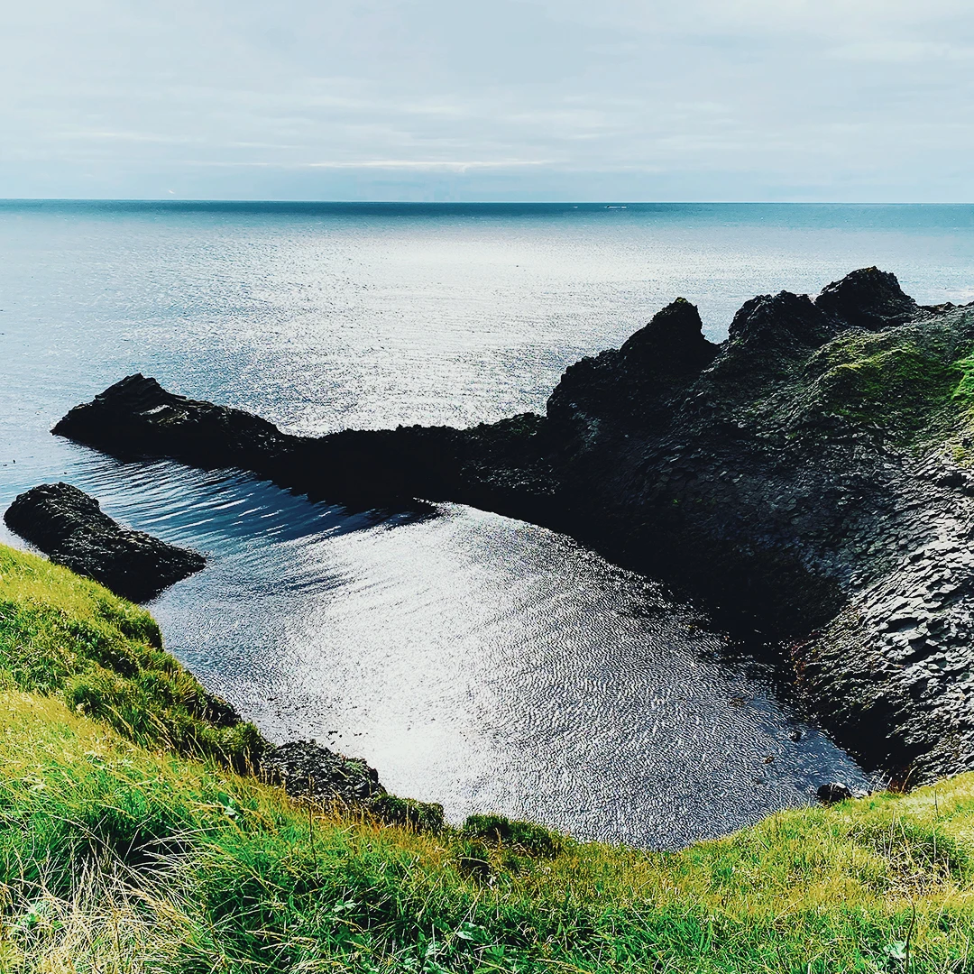 A small rocky coastal inlet with calm reflective water and green clifftops under a pale overcast sky