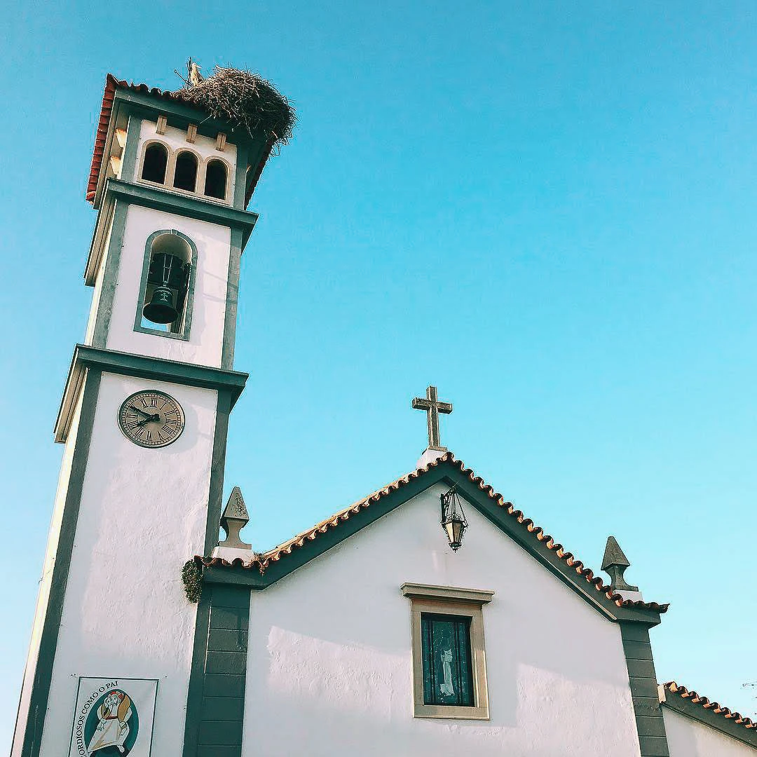 White Portuguese church with a green-trimmed clock tower topped by a stork nest against a bright blue sky