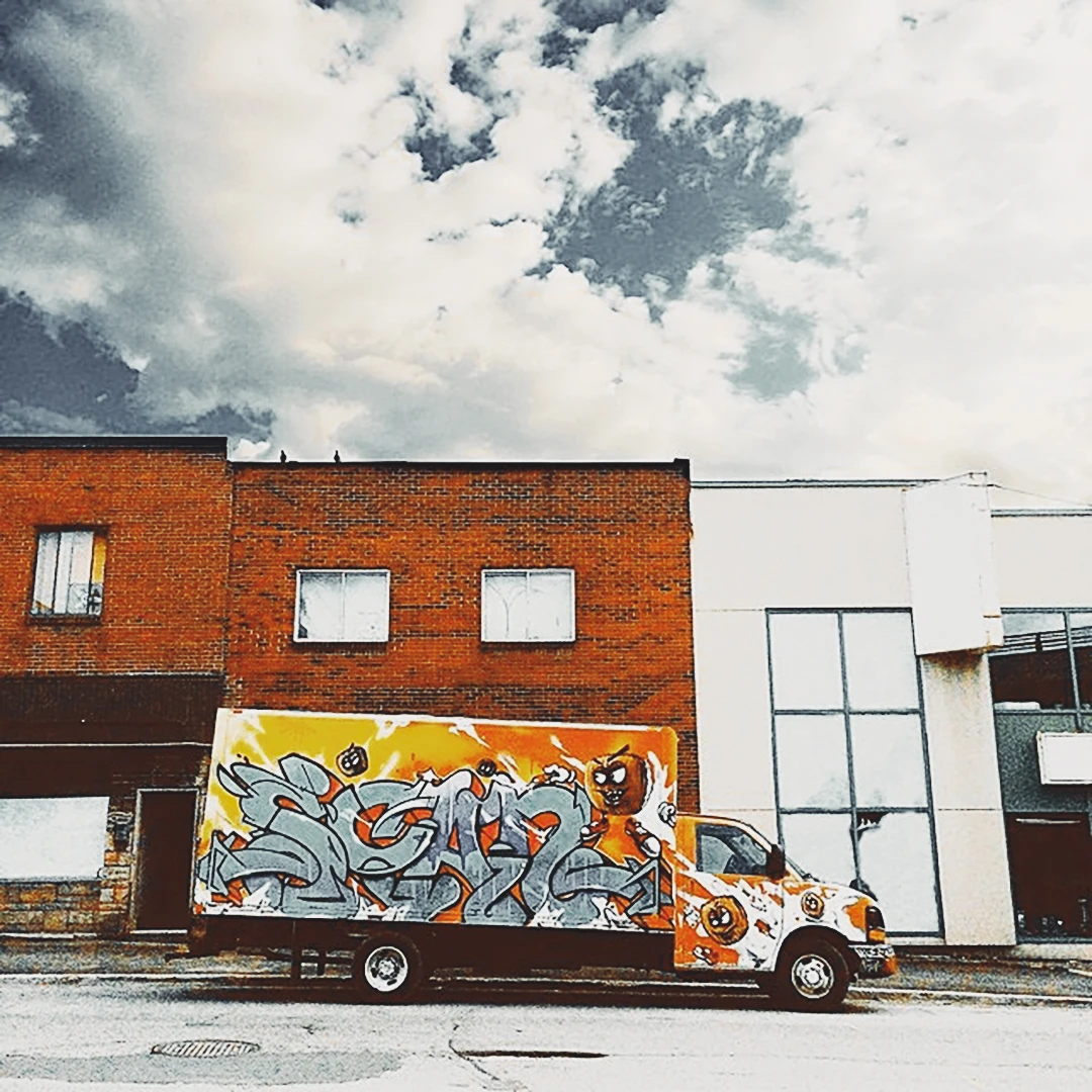 A delivery truck covered in bold orange and yellow graffiti parked outside a red brick building under dramatic clouds