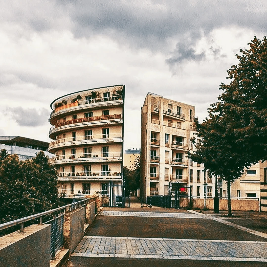 A curved modernist residential building with balconies on a quiet European street corner under a cloudy sky