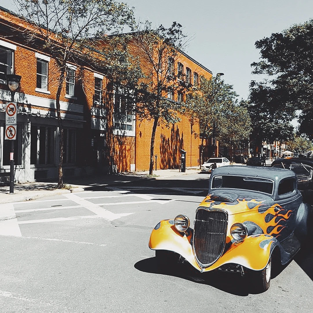A vintage hot rod with yellow flame paint job parked on a sun-drenched urban street lined with brick buildings