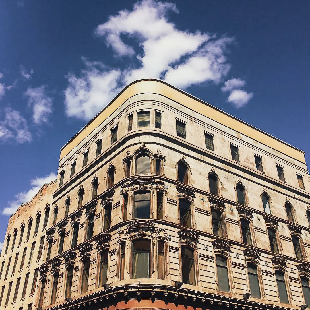 Looking up at the ornate stone corner facade of a Victorian-era commercial building against a bright blue sky with white clouds