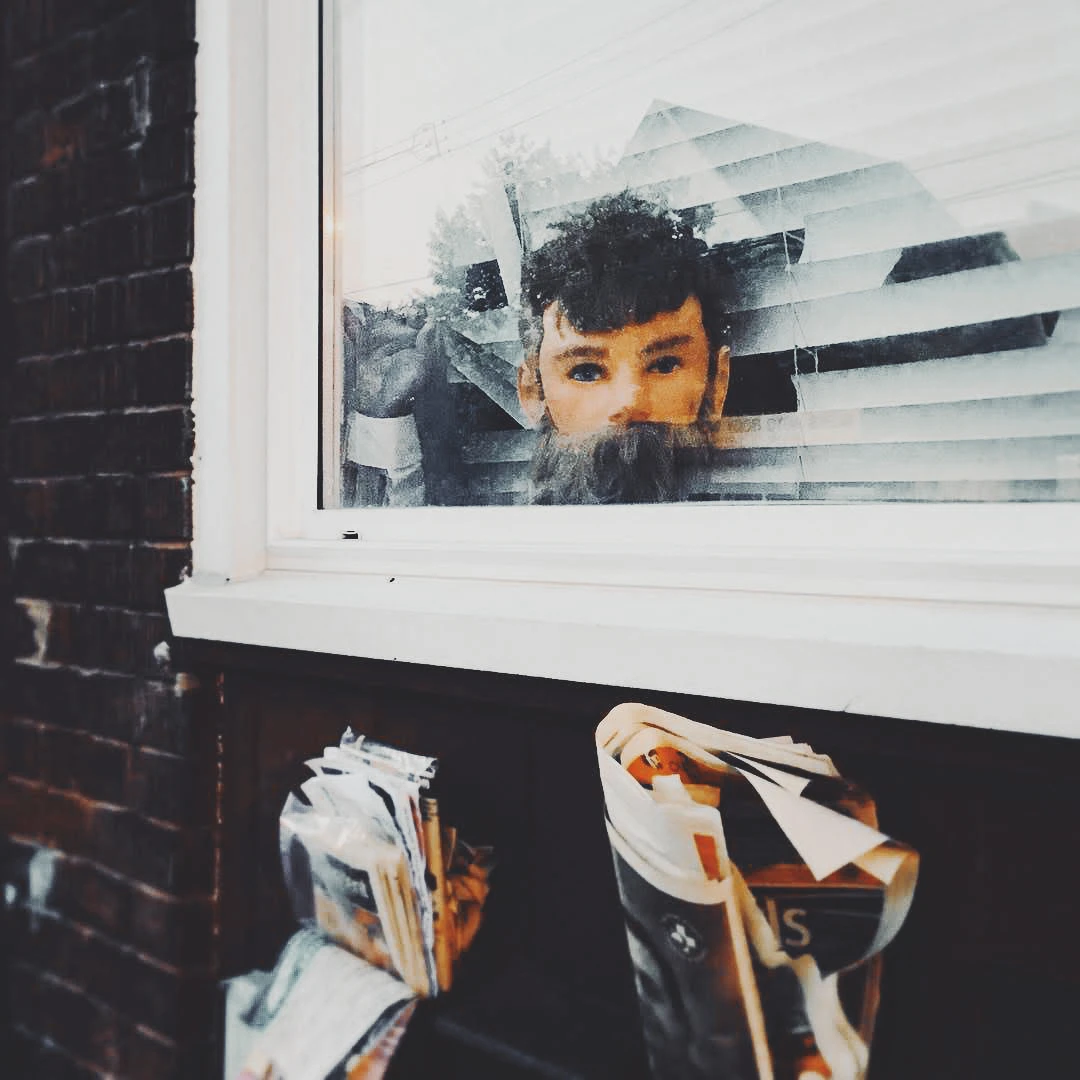 A bearded man peers curiously through a shop window, reflected in the glass alongside bundles of paper bags