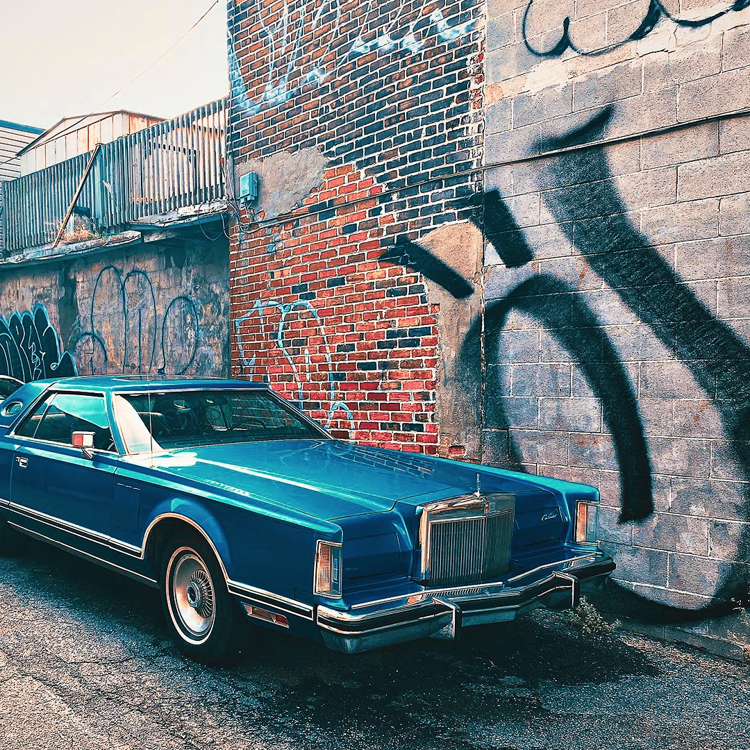 A classic blue Lincoln Continental parked in an alley beside a weathered brick wall covered in graffiti shadows