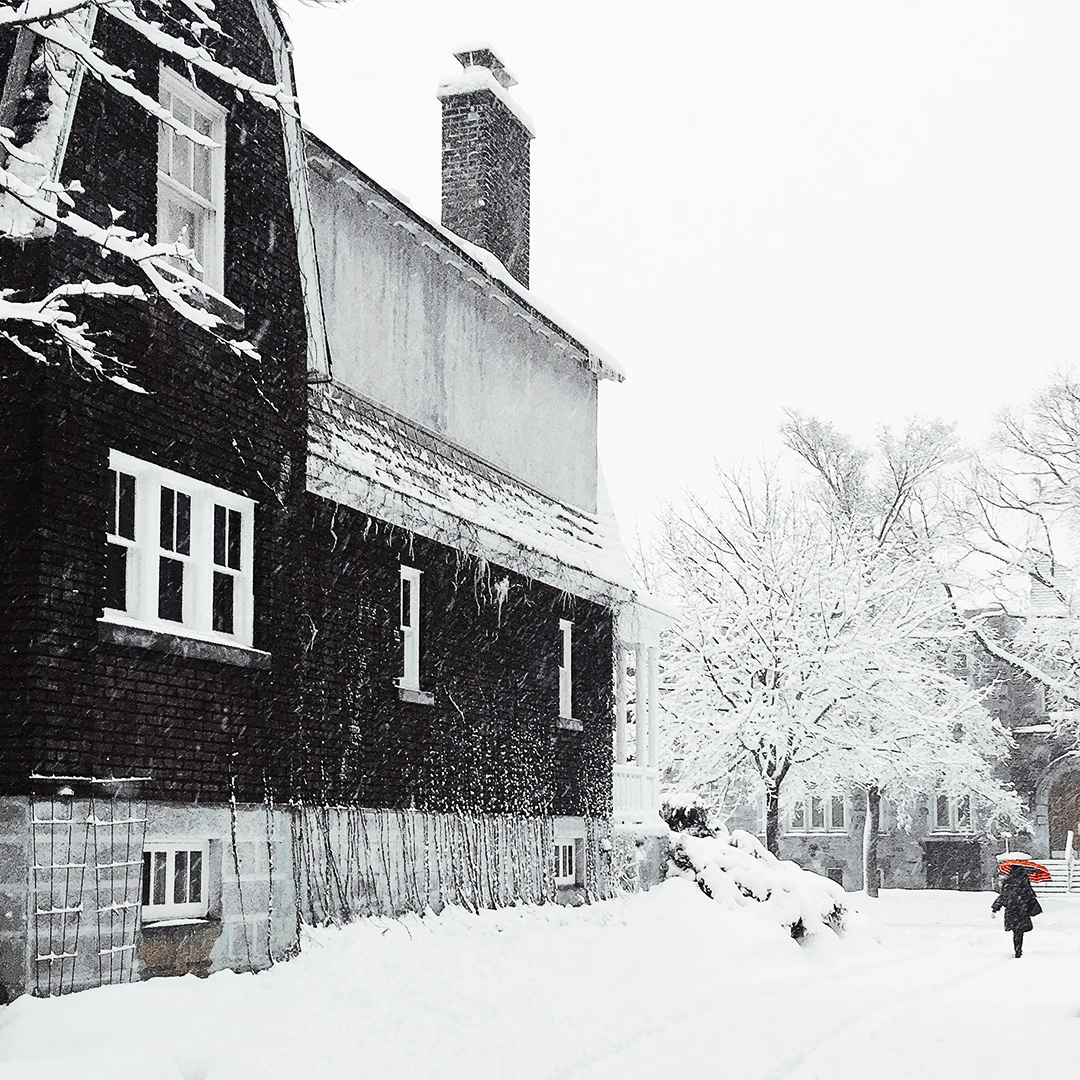 A lone figure with a red umbrella walks past a dark snow-blanketed house during a heavy winter snowfall