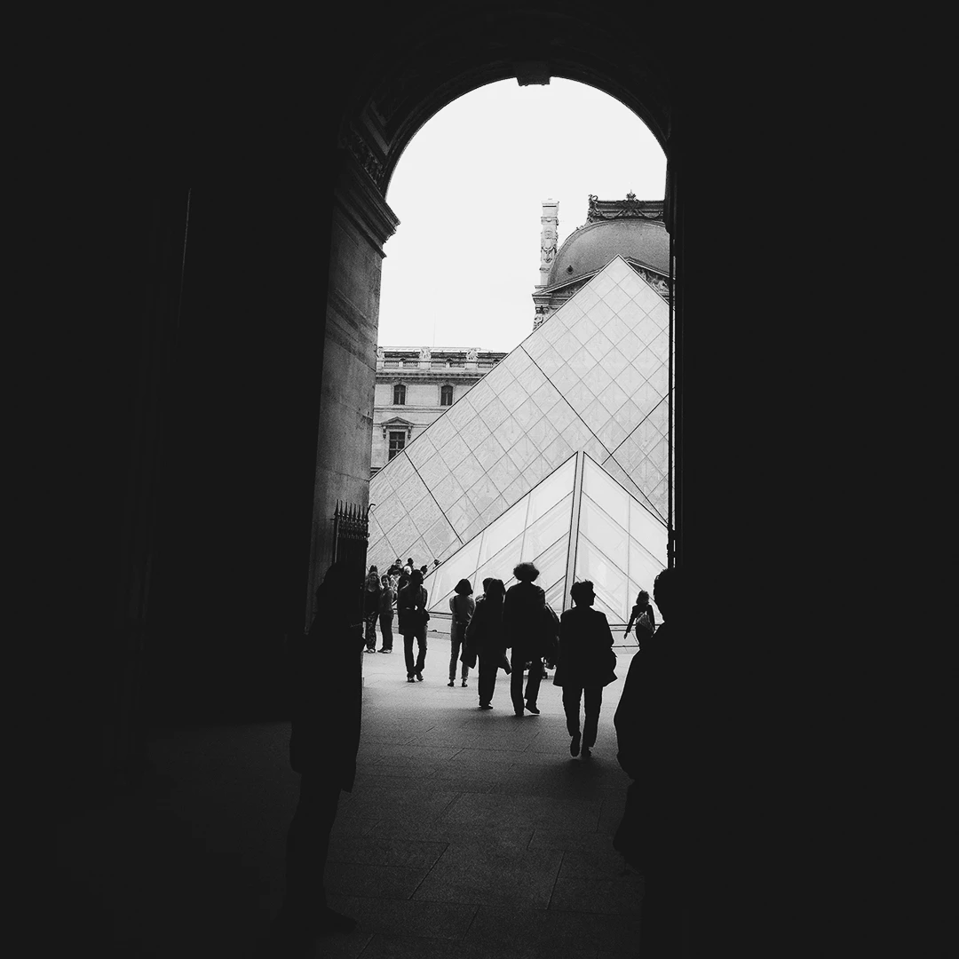 Black and white view of the Louvre Pyramid framed by a dark stone archway, silhouetted visitors crossing the courtyard