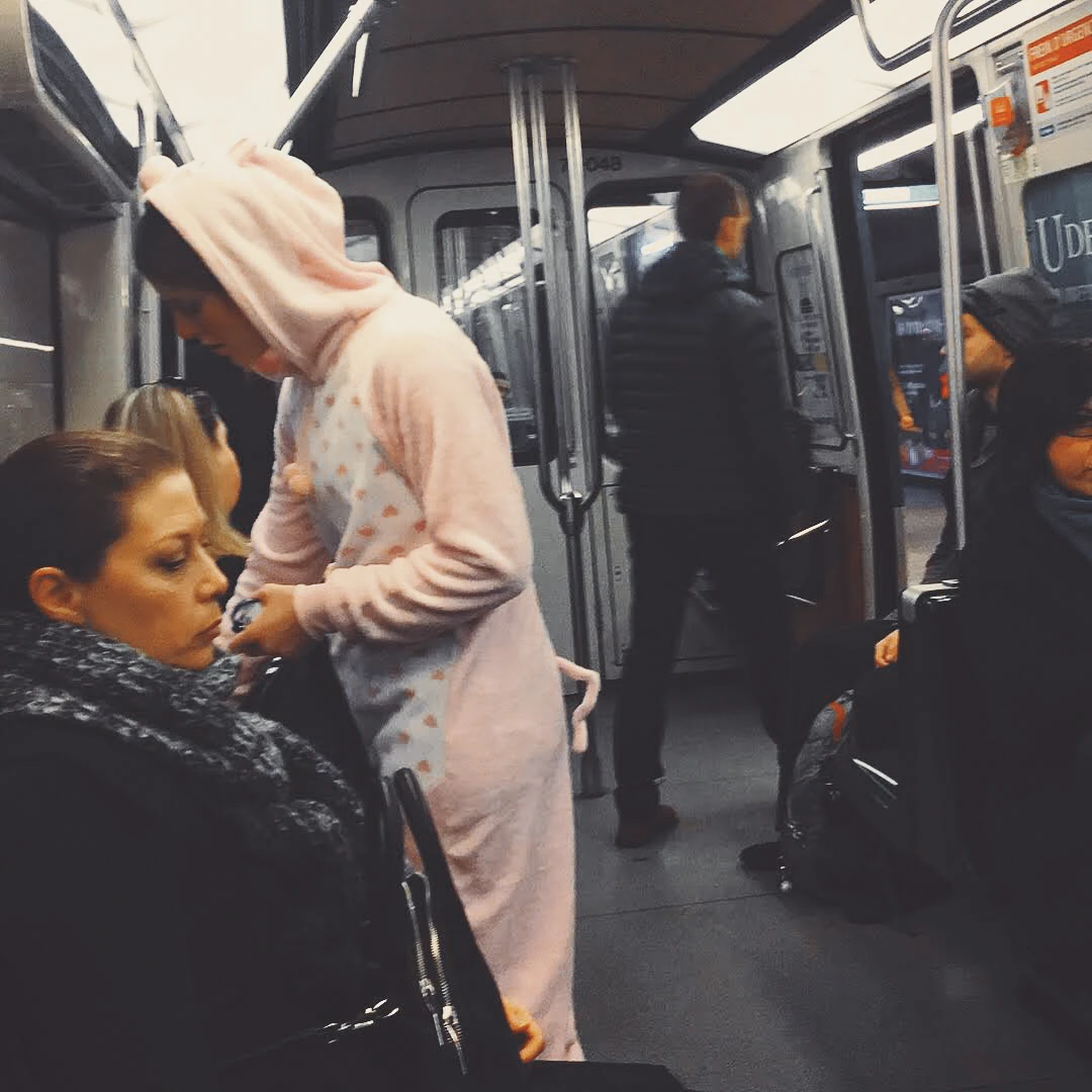 A person in a full pink unicorn onesie standing on a busy subway car surrounded by commuters in winter coats