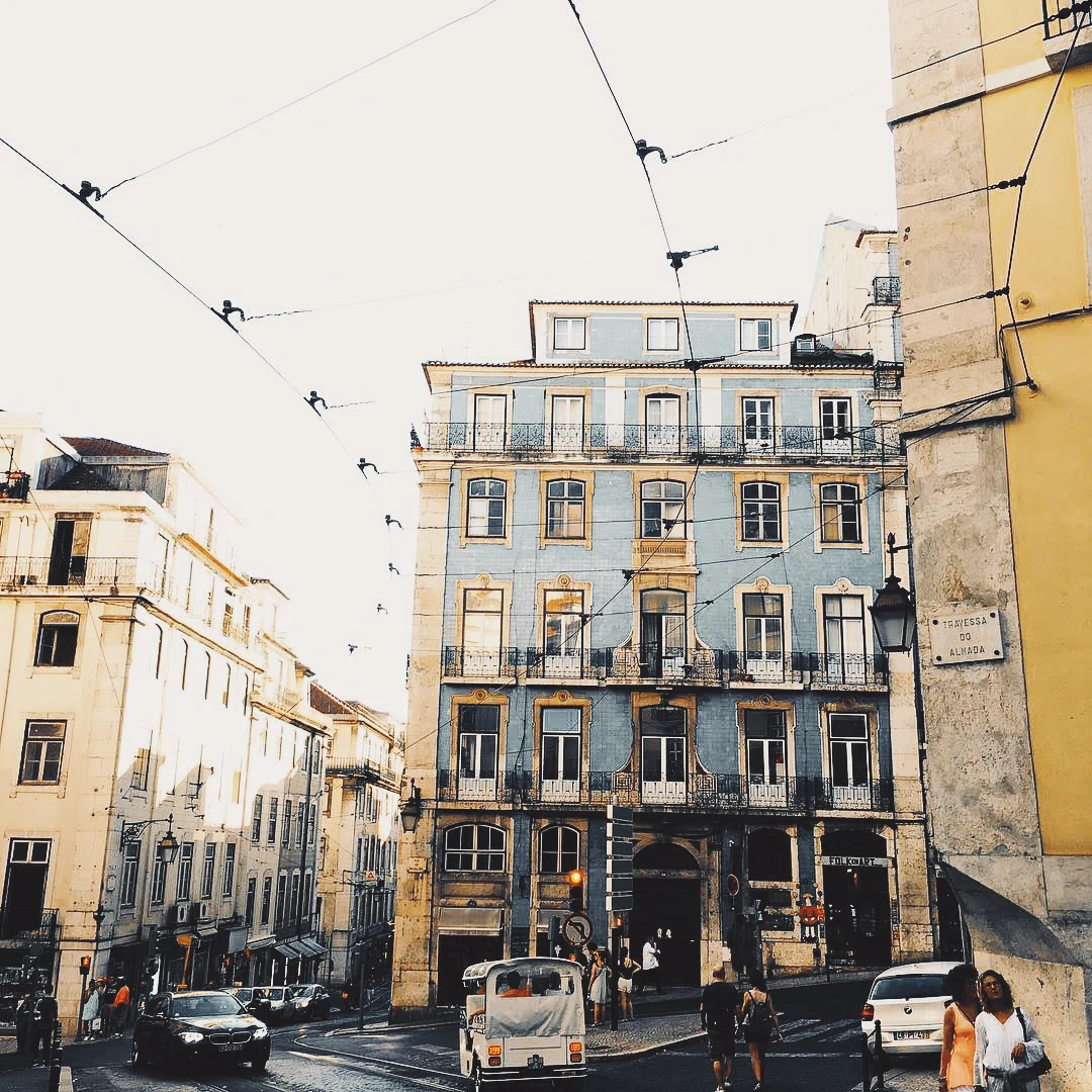 A faded blue tiled building at a Lisbon street corner, tram wires crisscrossing the pale sky above
