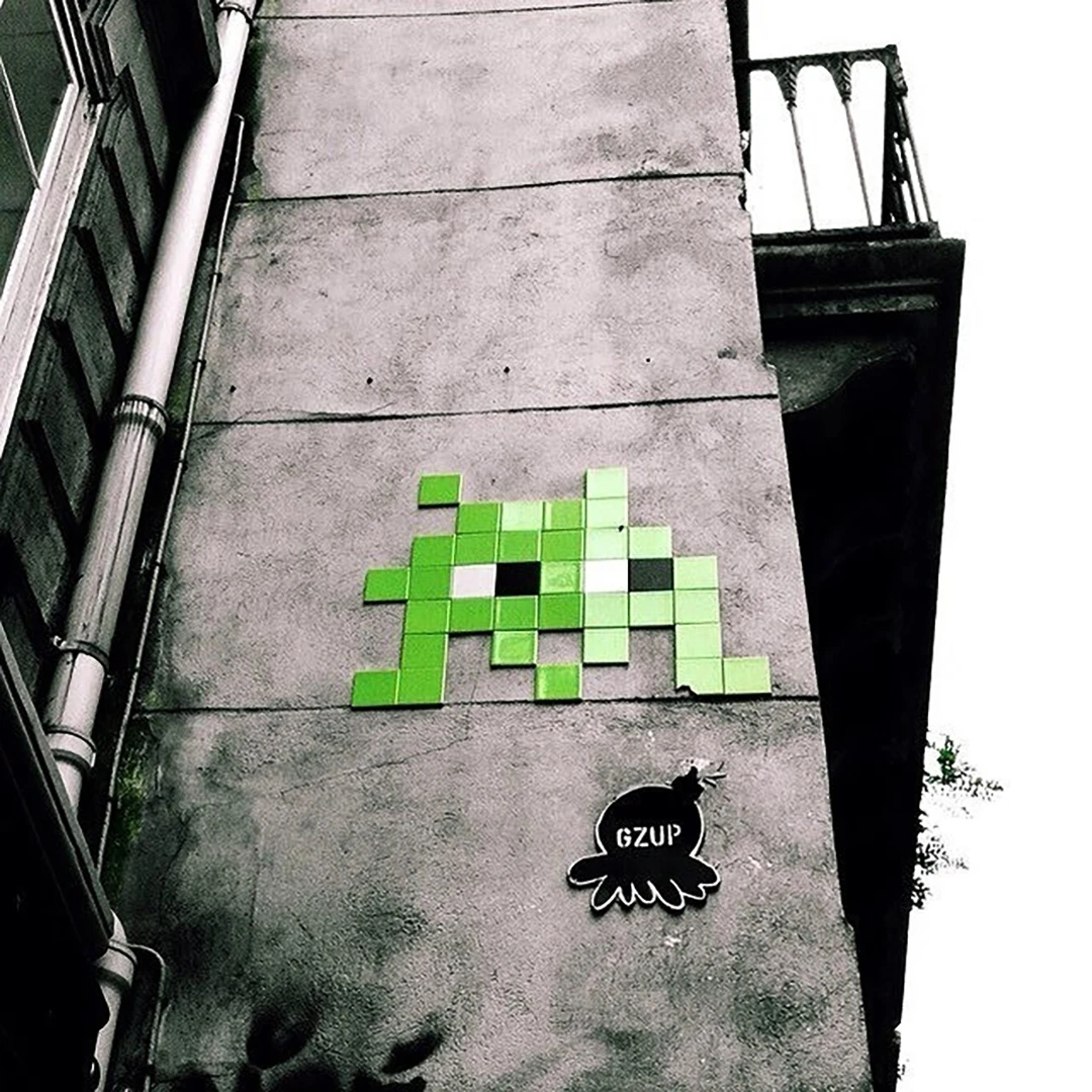 A green pixel Space Invader mosaic mounted on a concrete wall above a small crow sticker