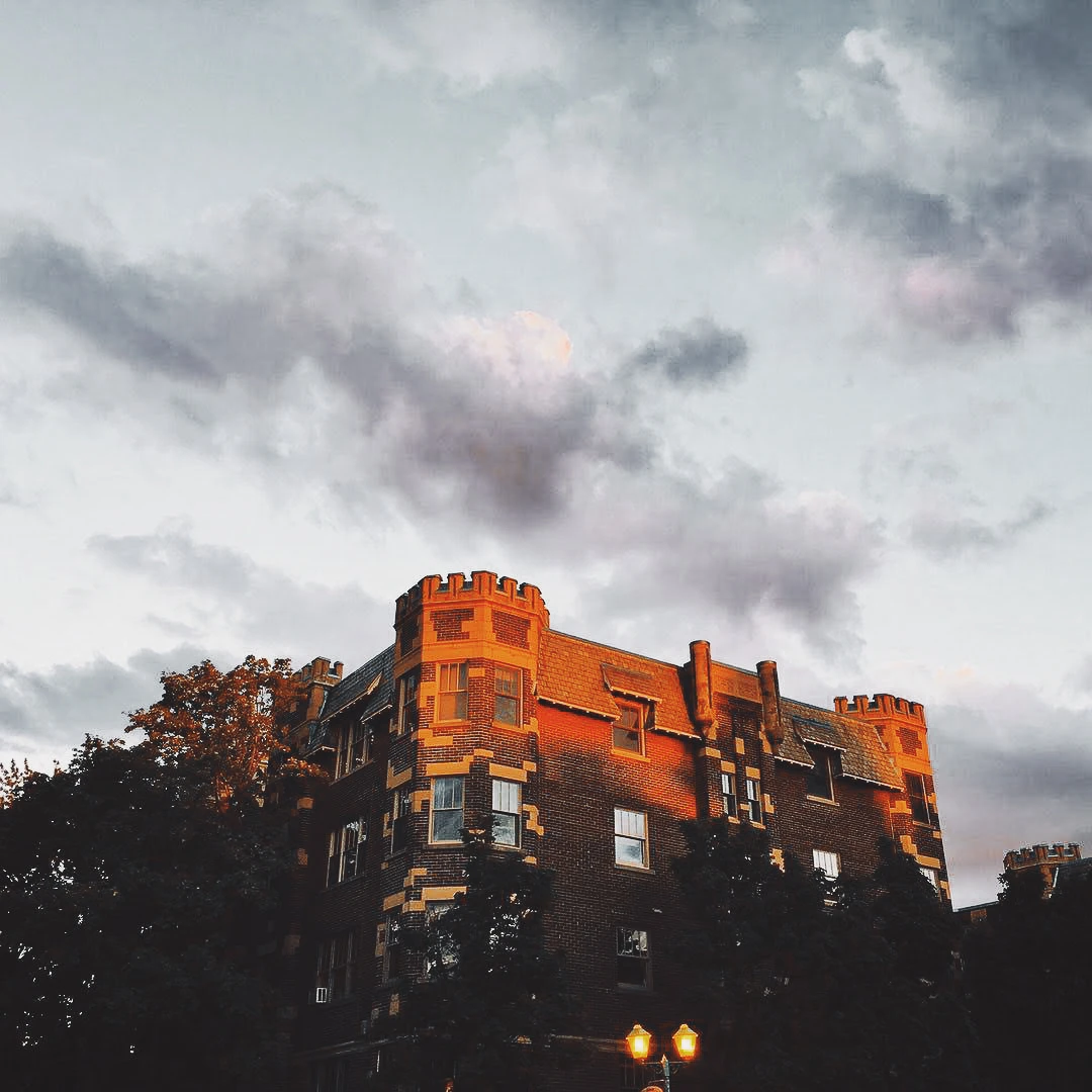 A dramatic red brick castle-like building with turrets glowing under a moody sunset sky with dark rolling clouds