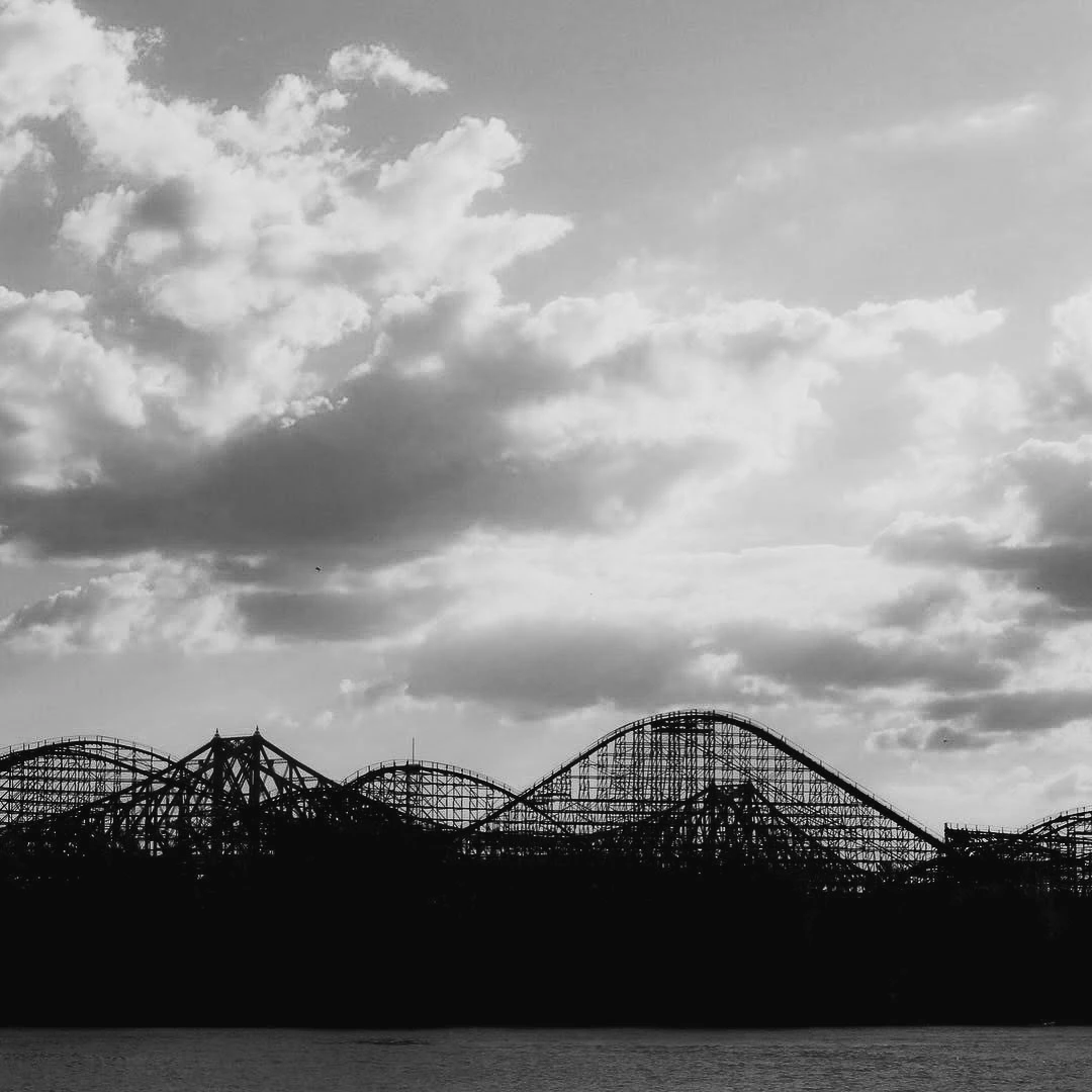 Black and white silhouette of a wooden roller coaster against a dramatic cloudy sky reflected in still water