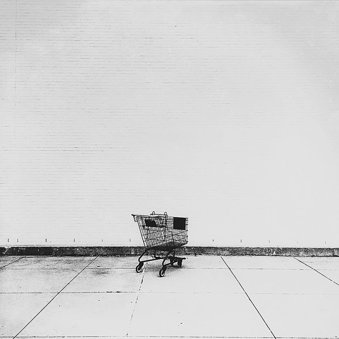 A lone shopping cart sits abandoned on a empty parking lot against a stark white brick wall