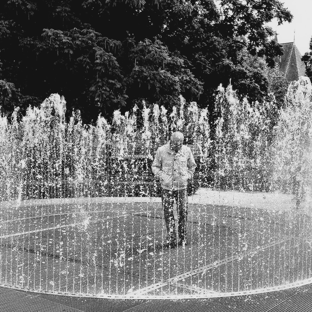 Black and white photo of an elderly man standing calmly inside a circular fountain with jets of water surrounding him