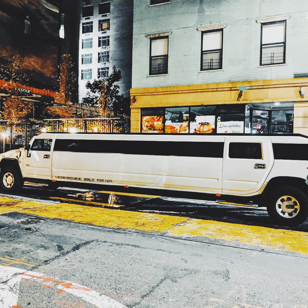 A white stretch Hummer limousine parked on a rain-slicked city street at night under yellow lights