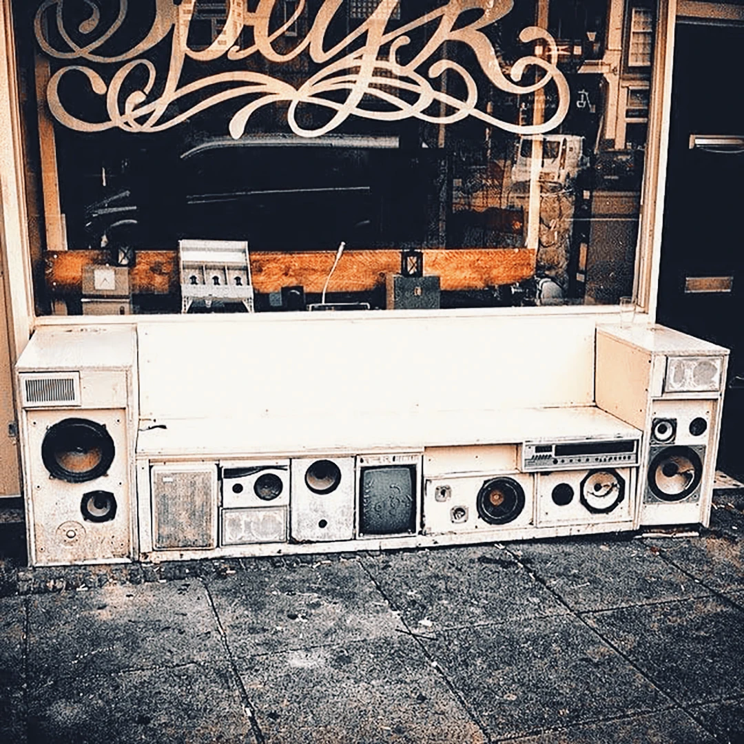 A bench made entirely of stacked vintage stereo speakers and radios sitting outside a shop with hand-lettered signage