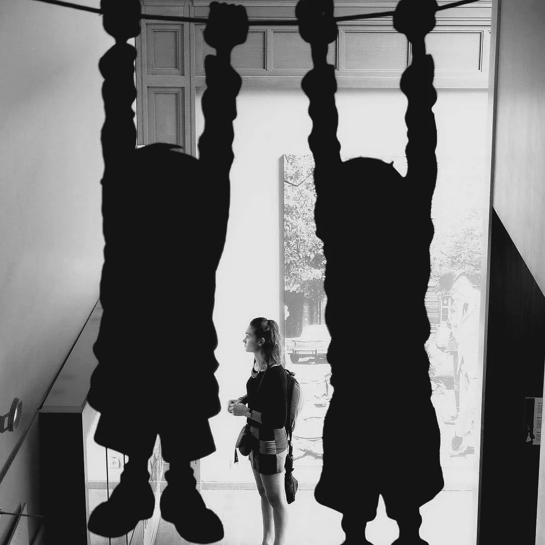Black and white photo of two inverted figure sculptures hanging from a ceiling, a woman standing between them looking toward a bright window