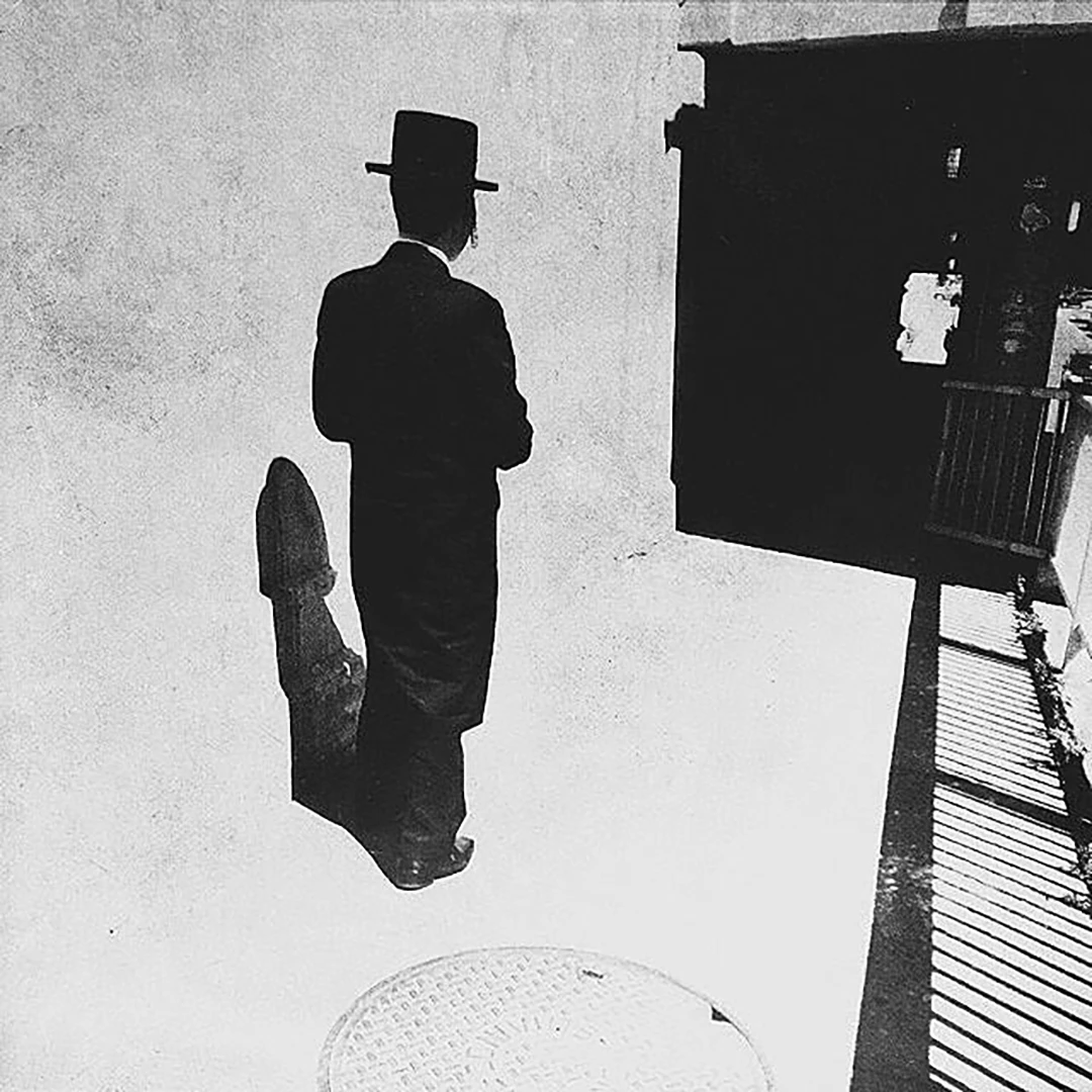 Black and white photo of a man in a top hat and long coat seen from behind, his sharp shadow stretching across a sunlit sidewalk