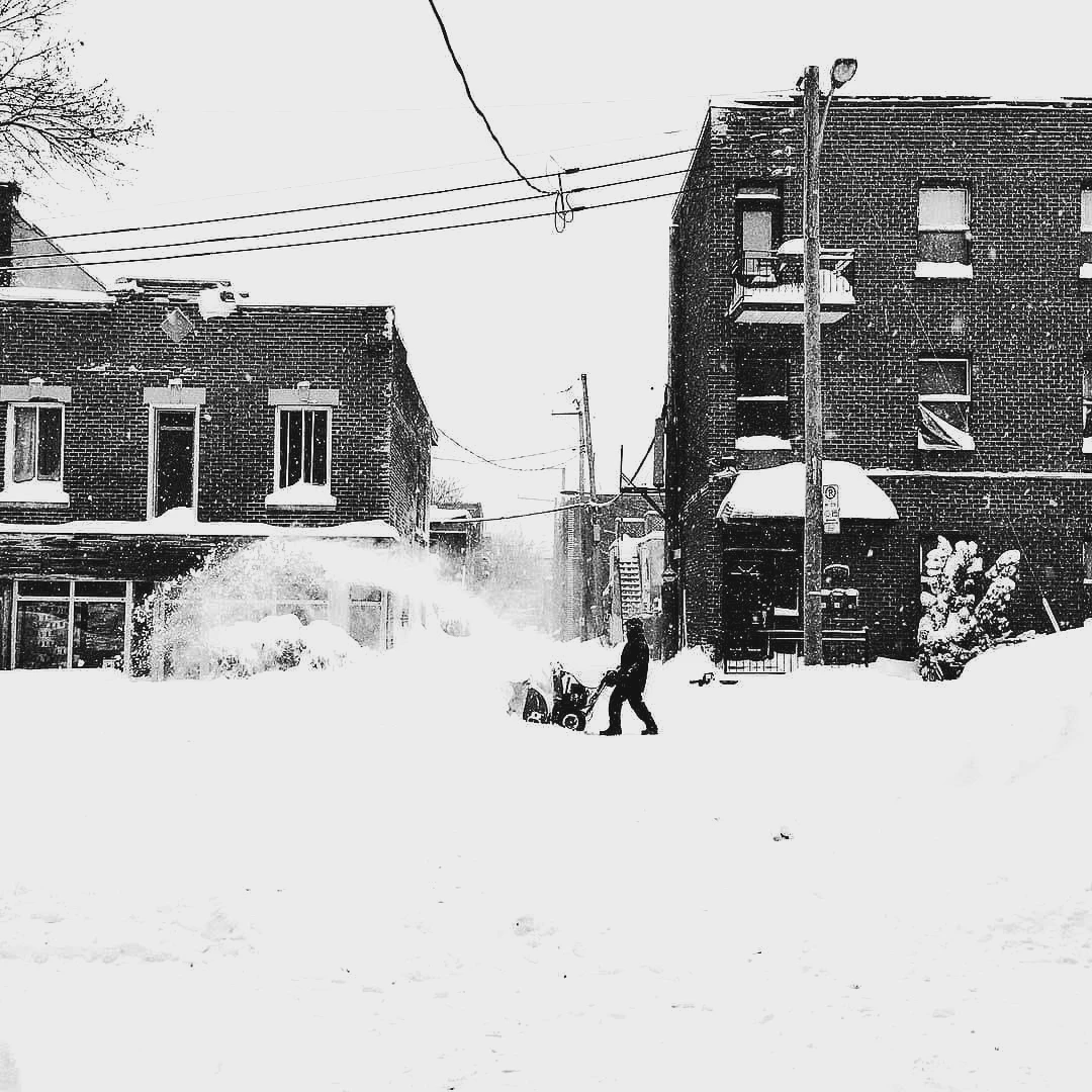 Black and white photo of a person using a snowblower on a completely snow-buried Montreal street during a blizzard