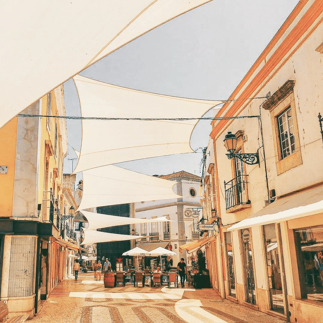 A sun-drenched pedestrian street in the Algarve covered by large shade sails, lined with cafes and colourful facades