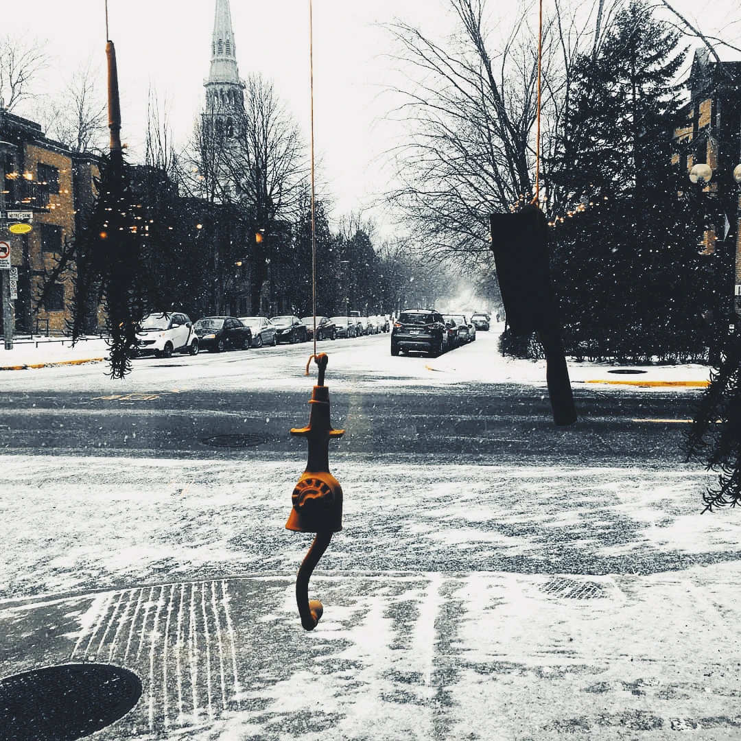 A small decorative figure hangs from a wire across a snowy Montreal street, a church steeple visible in the grey distance
