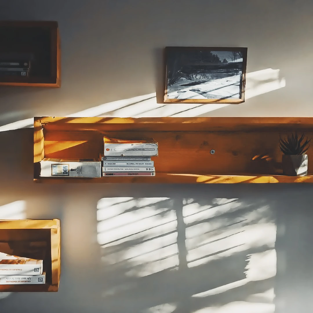 Warm afternoon sunlight casts long diagonal shadows across a wooden wall shelf holding books, cassettes, and a small framed photo