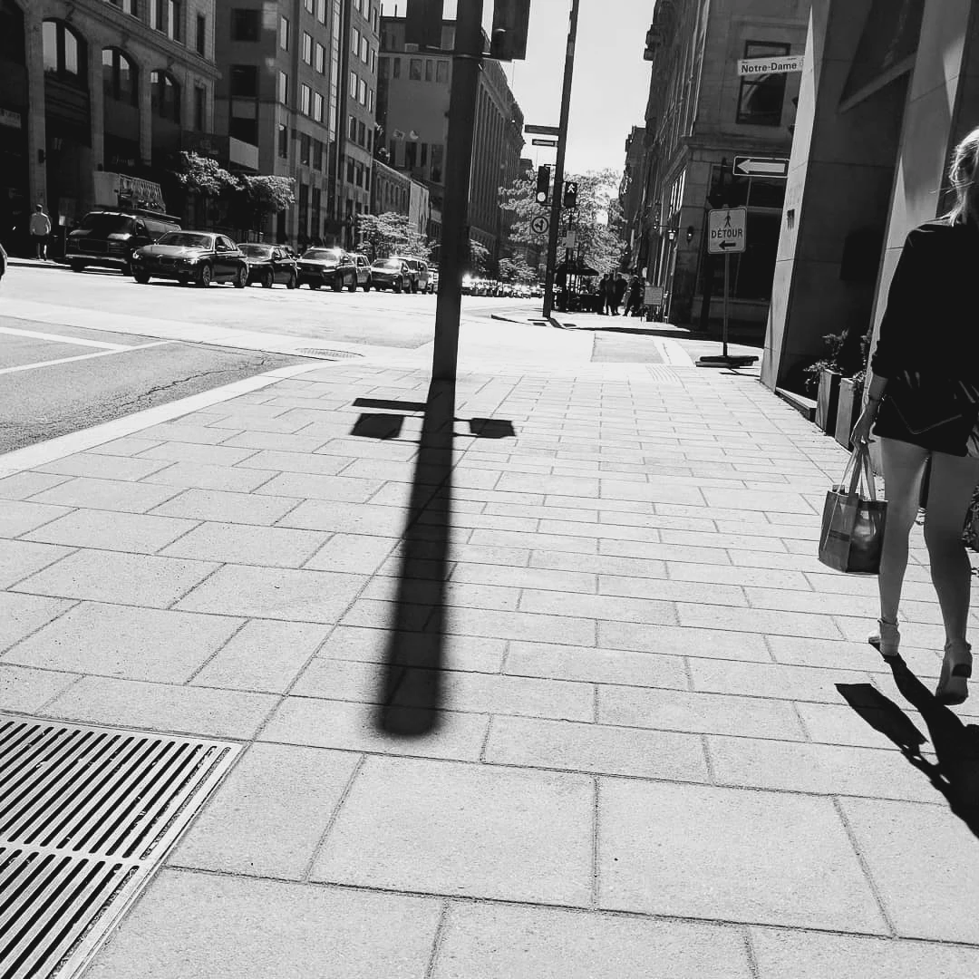 Black and white photo of a street pole casting an impossibly long shadow across a wide downtown sidewalk, a woman walking past