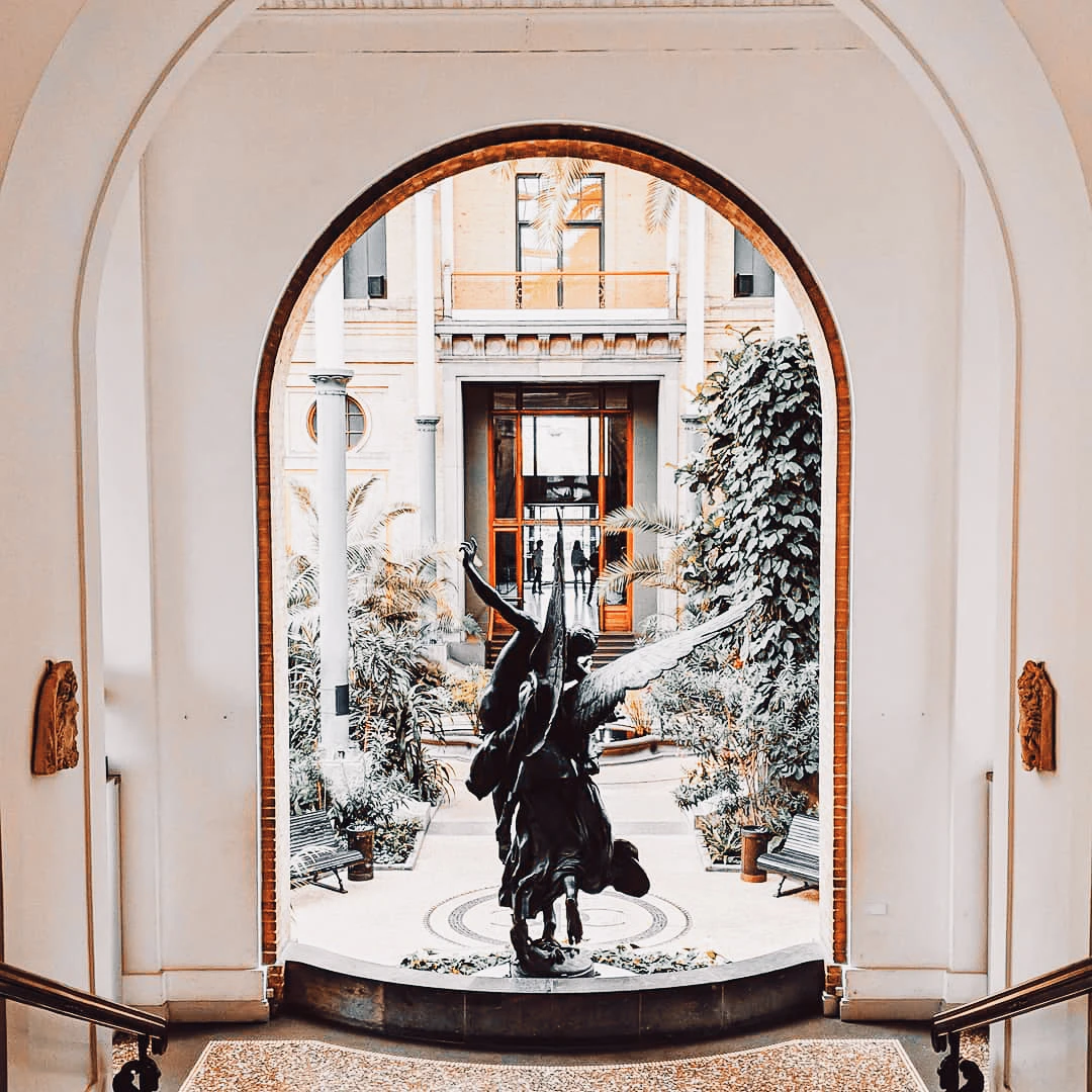 A dramatic black sculpture of a figure with outstretched wings stands at the centre of an elegant arched atrium filled with potted trees