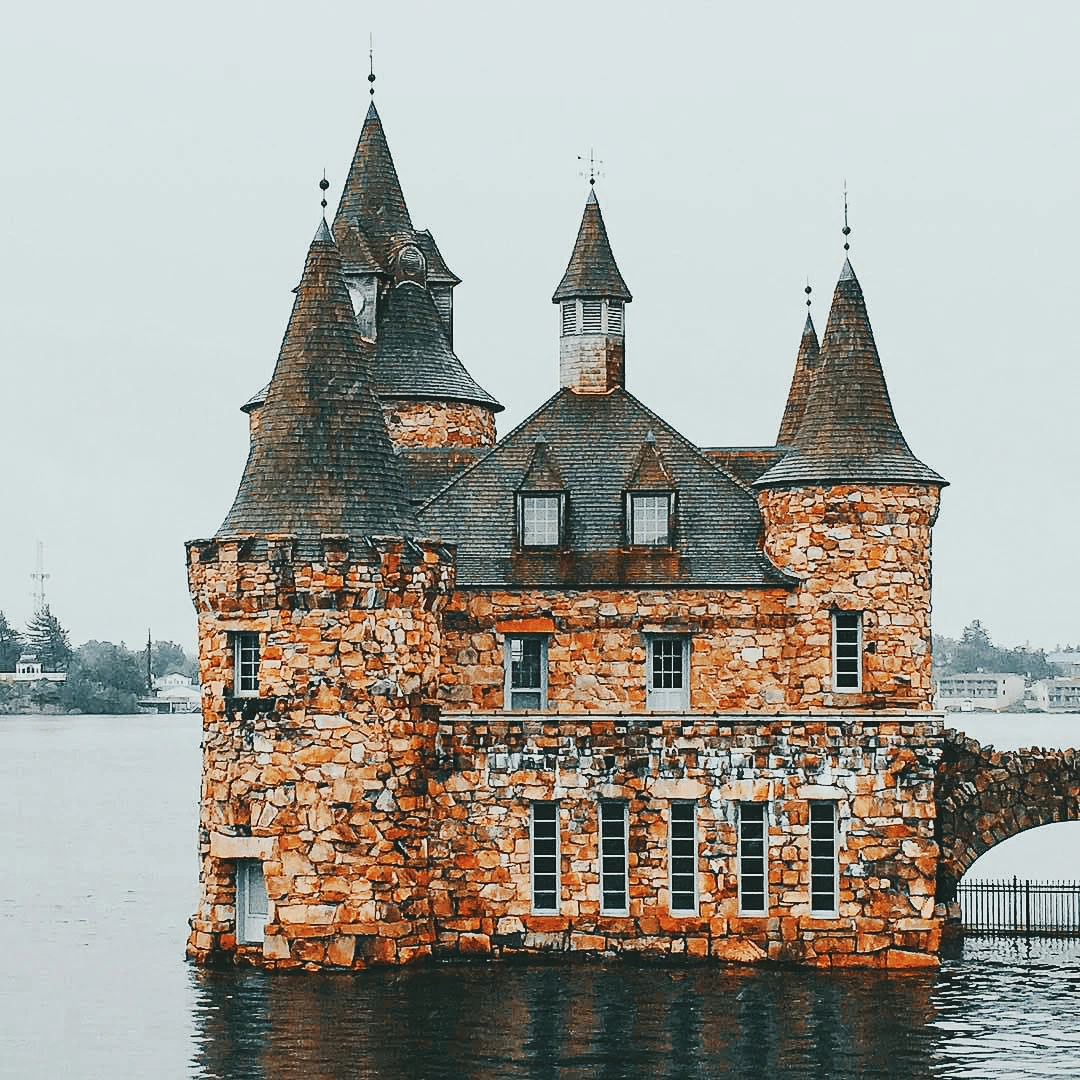 A stone castle tower rises from the middle of a grey river, its conical turrets and red brick walls reflected in the still water