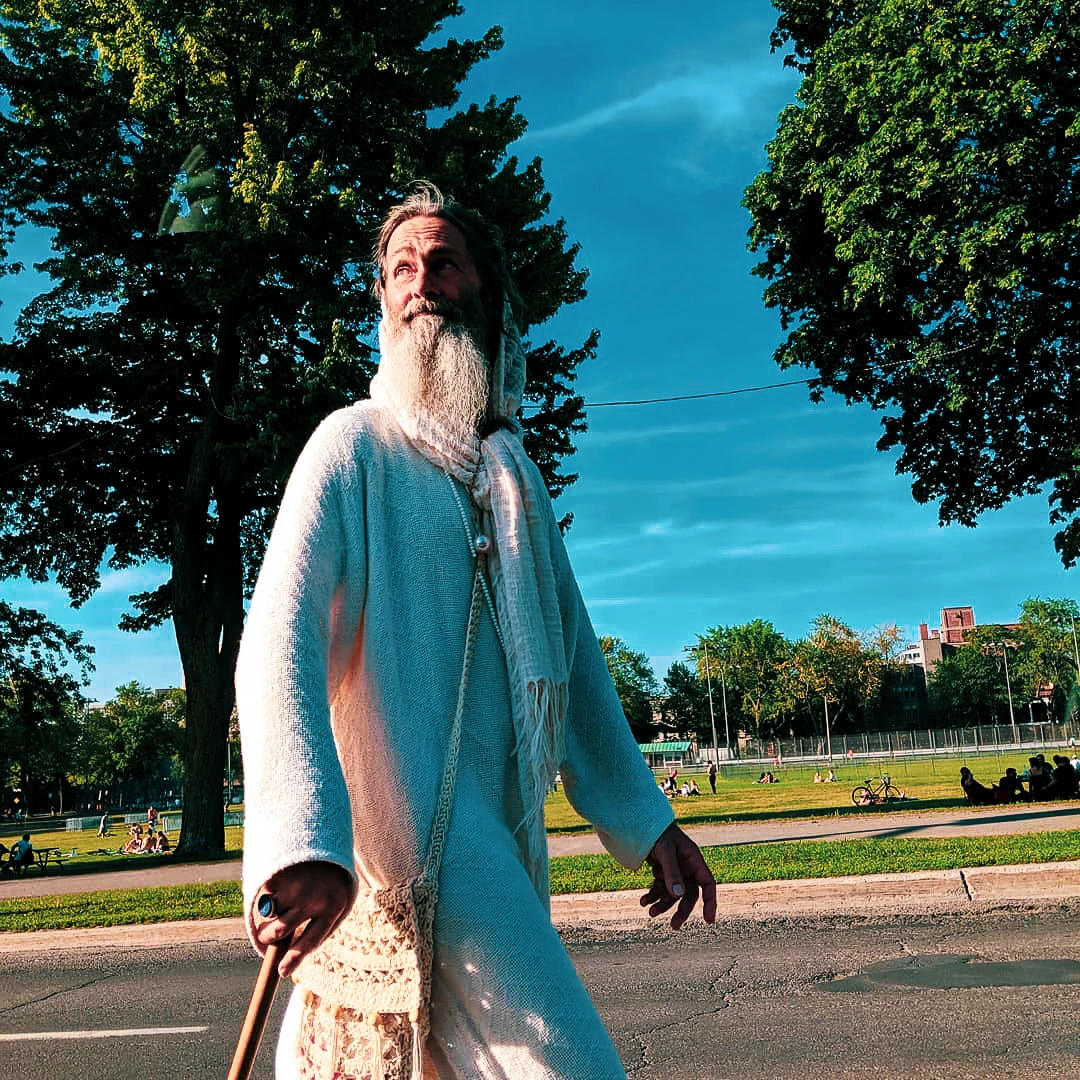 Man in a long white robe walking alone through a sunlit park with trees in the background