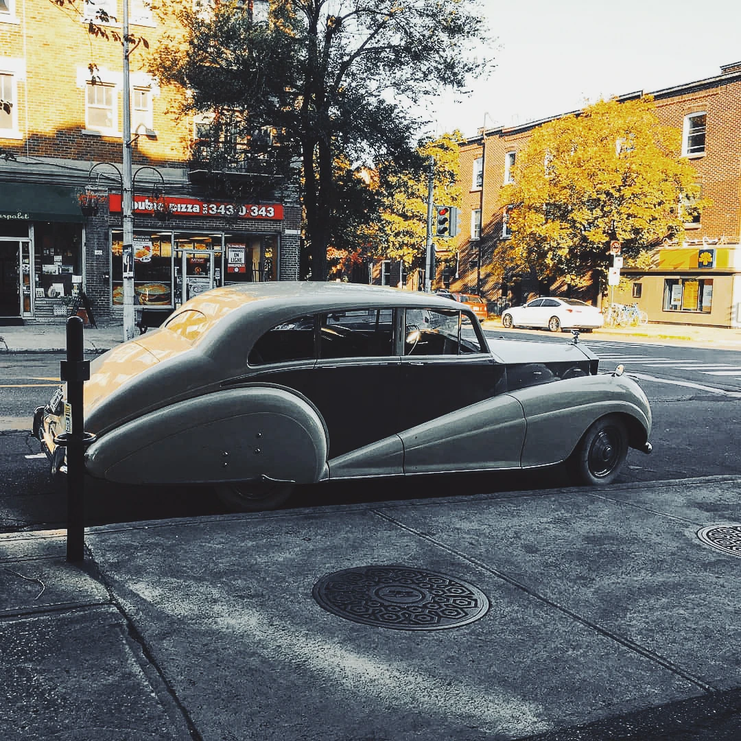 A vintage Rolls Royce-style luxury car parked on a sunny urban street lined with autumn trees