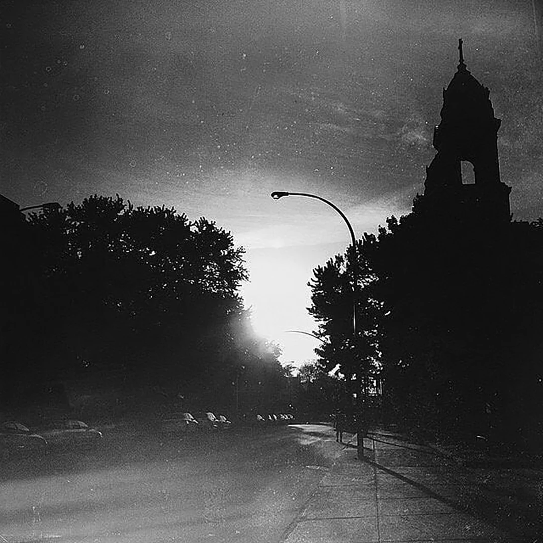 Black and white film-grain street scene with the silhouette of a church tower at dusk