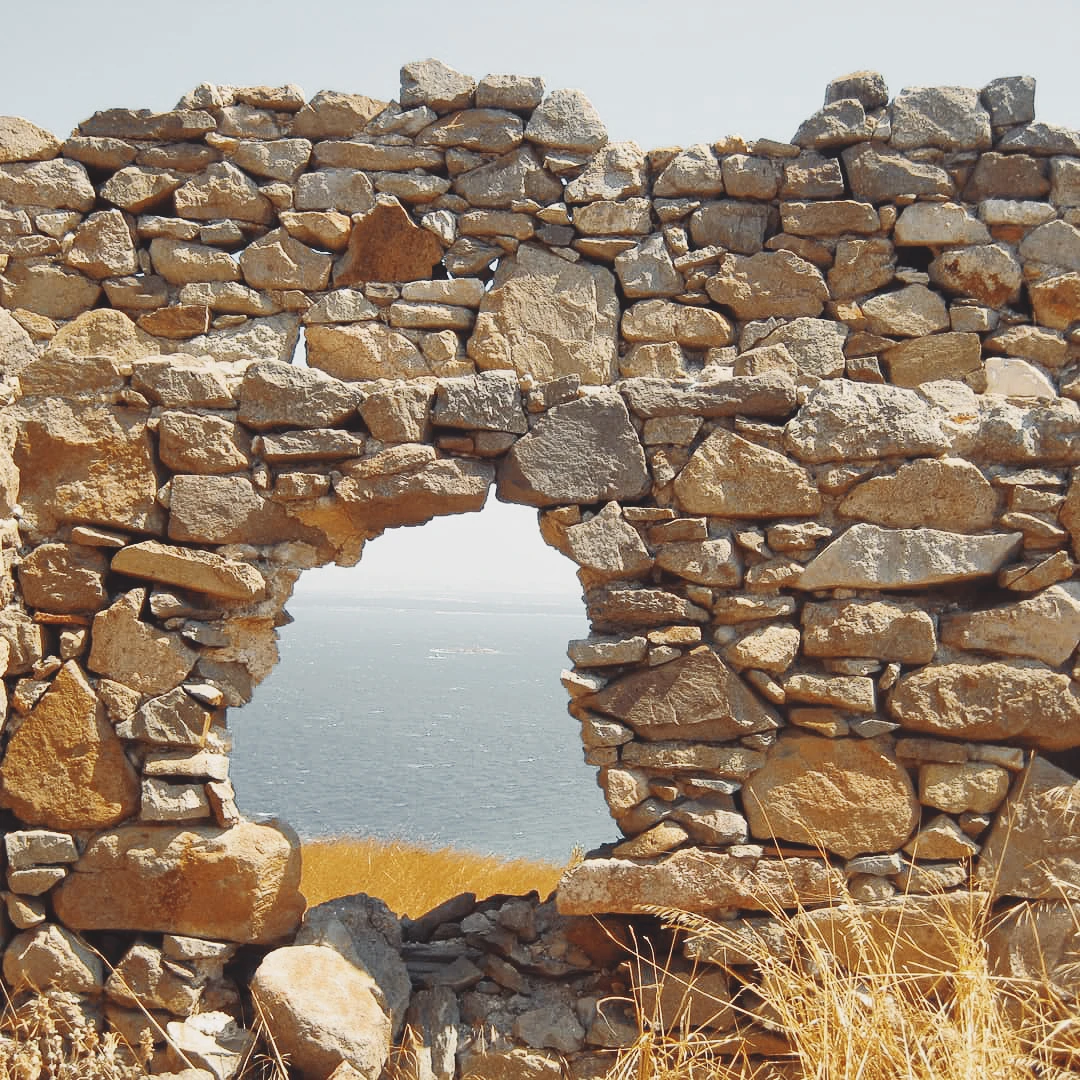 A round opening in an ancient stone wall framing a view of the sea beyond dry hillside