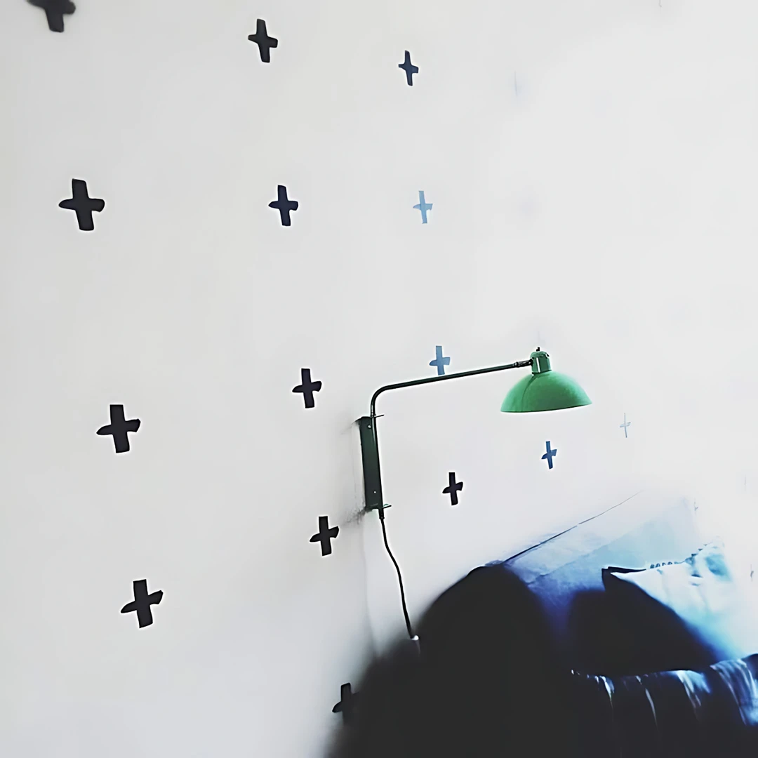 White bedroom wall covered in scattered black cross stickers beside a green swing-arm reading lamp