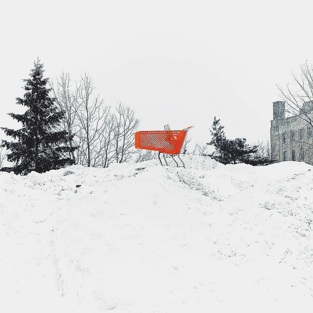 A lone orange shopping cart perched on top of a snow mound against a bleak winter sky