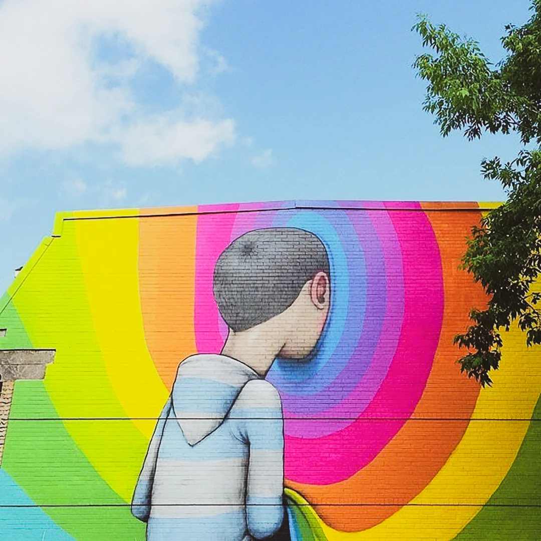 Large mural of a child seen from behind pressing their face into a radiating rainbow painted on a building wall